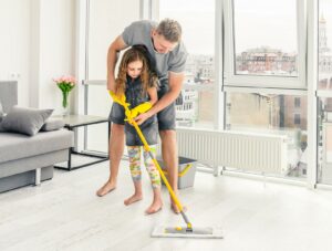 Ein Mann bezieht ein Mädchen in alltägliche Aktivitäten ein, indem er einen Boden in einem Wohnzimmer reinigt.
