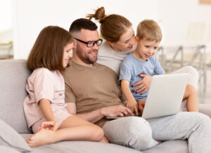 Eine vierköpfige Familie sitzt auf einem Sofa und schaut auf einen Laptop. Vater und kleiner Sohn spielen mit dem Laptop, während Mutter und Tochter lächelnd zuschauen und zeigen, wie alternsgerechte Mediennutzung Generationen durch Technologie zusammenbringen kann.