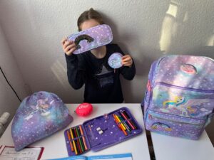 Welche Schultasche für mein Kind? Ein Kind hält eifrig ein Federmäppchen in der Hand, umgeben von lila Schulsachen mit Einhornmotiv, die verstreut auf dem Tisch liegen, während es einen Blick auf seine Schultasche wirft, die daneben steht.