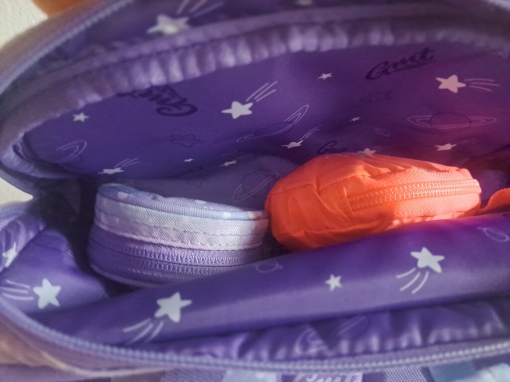 Offener Rucksack mit violetter Innenseite und Stern- und Planetenmuster sowie zwei Reißverschlusstaschen – eine lavendelfarbene und eine orangefarbene.