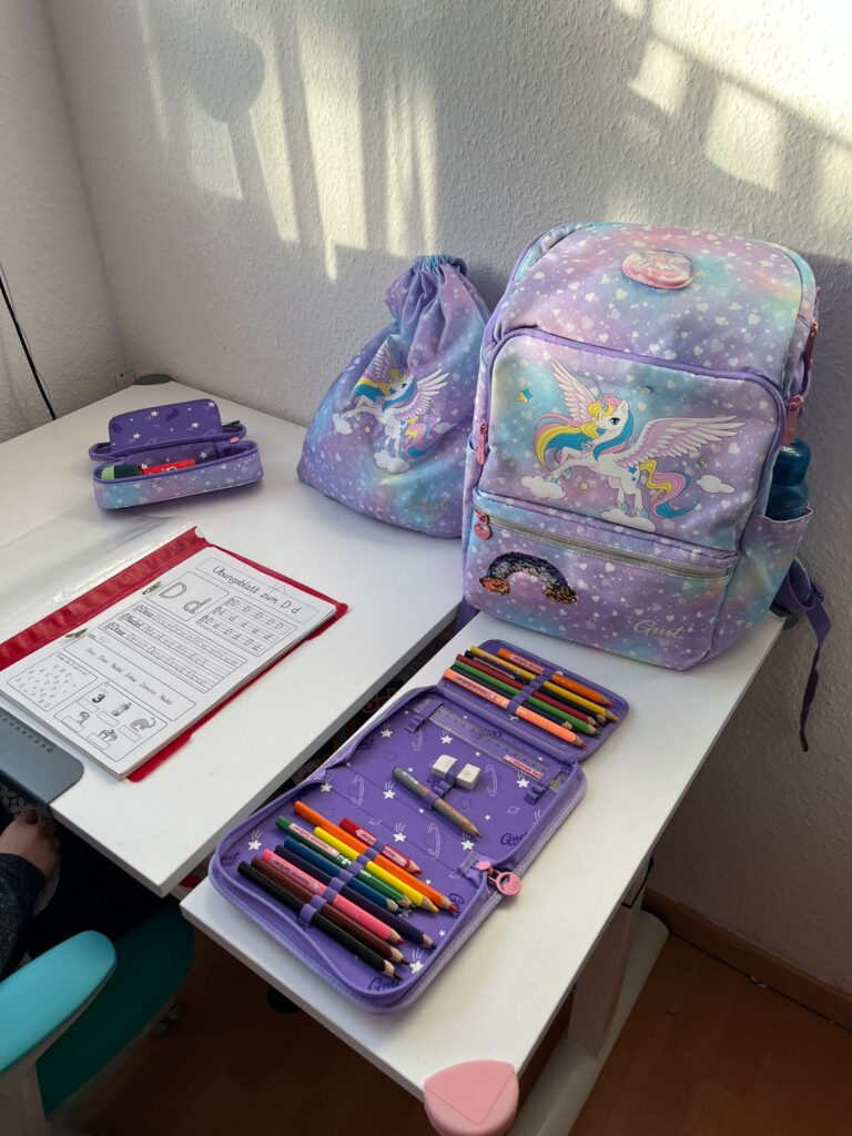 Ein Kinderschreibtisch mit einem lila Rucksack, einem Federmäppchen und einem Beutel mit Kordelzug und Einhornmotiven. Auf dem Schreibtisch liegt auch ein offenes Notizbuch mit Aufschrift.