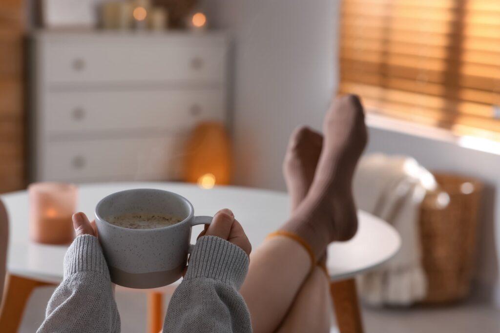 Eine Zeit zu entspannen kann deine tägliche Gelassenheit fördern. Eine Person genießt einen Moment der *Zeit zum Entspannen*, hält eine Kaffeetasse in der Hand und hat ihre Füße bequem auf dem Tisch abgelegt. Die warme Beleuchtung, das sanfte Leuchten der Kerze, der gemütliche Korb und die weichen Jalousien im Hintergrund schaffen eine Atmosphäre vollkommener Entspannung.