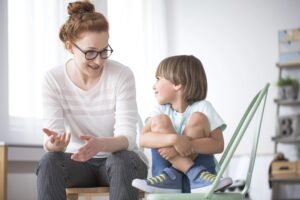 Kindern Liebevoll Grenzen setzten. In einem hellen Raum sitzt eine Frau mit Brille auf einem grünen Stuhl und erklärt einem kleinen Jungen behutsam Grenzen.